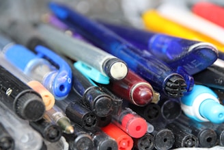 A close-up of a scattered collection of various pens, including ballpoint pens and highlighters. The pens are of different colors, primarily featuring blue, black, red, and yellow. Some pens have transparent bodies, revealing the ink inside, while others are opaque. The collection appears to be a mix of new and used pens, arranged haphazardly.