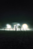 A dimly lit soccer field at night is enveloped in mist, creating a mysterious and atmospheric scene. The foreground features a soccer goal and railings under the glow of floodlights, casting a soft, diffused light that highlights the fog.