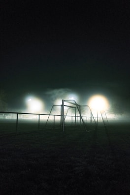 A dimly lit soccer field at night is enveloped in mist, creating a mysterious and atmospheric scene. The foreground features a soccer goal and railings under the glow of floodlights, casting a soft, diffused light that highlights the fog.