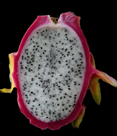 A close-up view of a sliced dragon fruit, revealing its white flesh speckled with small black seeds. The vibrant pink rim of the fruit contrasts sharply with the dark background, highlighting the fruit's unique appearance.