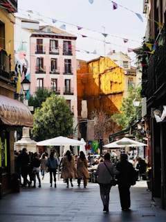 A bustling urban street lined with shops and cafes, featuring colorful buildings. People are walking and sitting under umbrellas, enjoying the lively atmosphere. Above, strings of triangular flags add a festive touch.