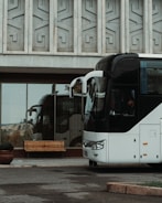 A large white bus is parked in front of a building with a patterned concrete facade. Reflections of the bus and surrounding area can be seen in the large glass windows of the building. A wooden bench is positioned near the entrance, and there is a potted plant beside it. The driver is visible inside the bus.