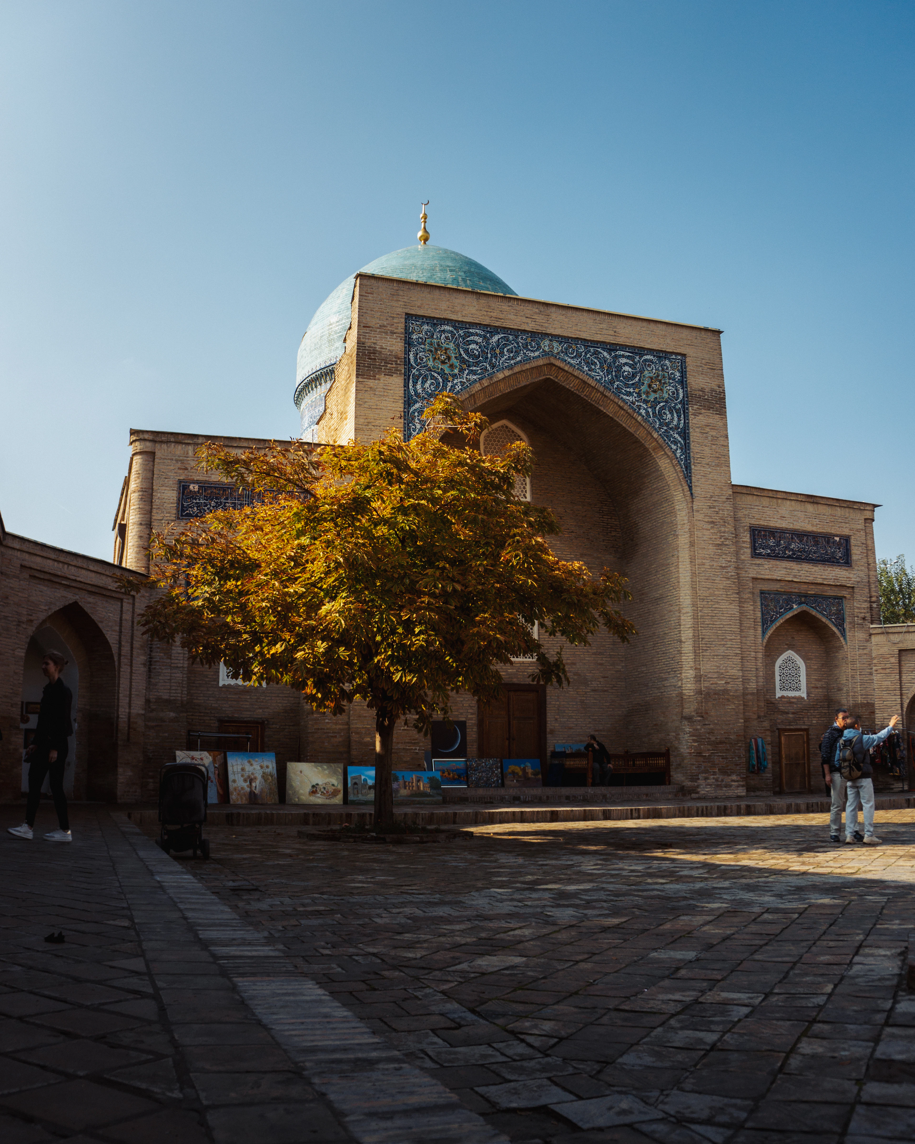 A tiled madrasa portal in Tashkent with a turquoise dome behind it and an autumn tree in the courtyard