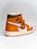 A stylish athletic shoe with a high-top design, featuring a combination of orange, brown, and white colors. The shoe displays a prominent logo on the side and a tag attached to the laces.