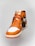 A close-up view of an orange and white high-top athletic shoe with a Nike Air logo on the tongue and orange laces. The shoe has a prominent leather texture and features a brown tag attached to one lace. The sole is orange, contrasting with the white front.