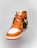 A close-up view of an orange and white high-top athletic shoe with a Nike Air logo on the tongue and orange laces. The shoe has a prominent leather texture and features a brown tag attached to one lace. The sole is orange, contrasting with the white front.