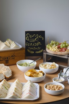 An assortment of food items is arranged on a wooden table. There are triangular sandwiches stacked on a platter, bowls filled with guacamole, tortilla chips, potato chips, and a mix of nuts. A blackboard with handwritten text in Spanish is positioned in the background. A rustic wooden tray contains skewers of grapes and other fruits.