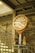 An ornate clock with Roman numerals is mounted on a stand in a rustic, industrial setting. The background features a brick wall and a flight of stairs. Two paper shopping bags are placed beneath the clock on a small table.