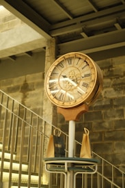 An ornate clock with Roman numerals is mounted on a stand in a rustic, industrial setting. The background features a brick wall and a flight of stairs. Two paper shopping bags are placed beneath the clock on a small table.