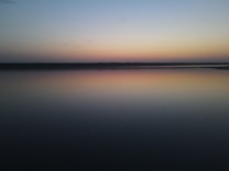 A tranquil and serene lake at sunset, with calm water reflecting the soft pastel shades of the sky. The horizon is a smooth, dark line, and the sky transitions from light orange to deep blue.