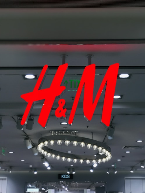 Bright red logo of H&M hangs in a retail store interior, surrounded by modern circular lighting fixtures on the ceiling. The store has a contemporary design with a variety of ceiling lights.