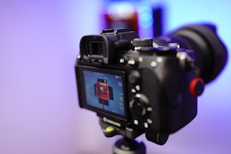 A camera is mounted on a tripod with the LCD screen displaying a preview of a red rectangular object. The background is blurred with cool blue and purple tones.