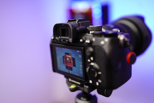 A camera is mounted on a tripod with the LCD screen displaying a preview of a red rectangular object. The background is blurred with cool blue and purple tones.