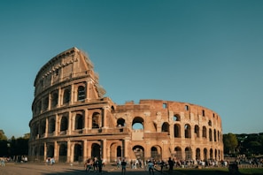 The Colosseum in Rome is bathed in warm sunlight, highlighting its ancient stone architecture with its series of arches and columns. A clear blue sky forms the backdrop, while tourists and visitors can be seen walking around the historic site.