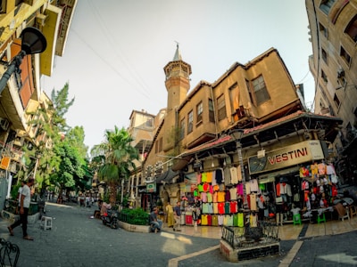 A bustling street scene with historic buildings, showcasing a shop displaying vibrant clothing. The architecture is traditional with a tower rising above the stores. People are walking and sitting around, adding to the lively atmosphere. Trees and greenery add a touch of nature amidst the urban setting.