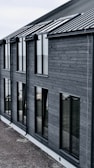 A modern building with dark wooden cladding and large rectangular windows. The roof is sloped and made of dark metal. The overall design is simple and minimalist.