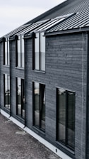 A modern building with dark wooden cladding and large rectangular windows. The roof is sloped and made of dark metal. The overall design is simple and minimalist.