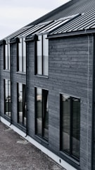 A modern building with dark wooden cladding and large rectangular windows. The roof is sloped and made of dark metal. The overall design is simple and minimalist.
