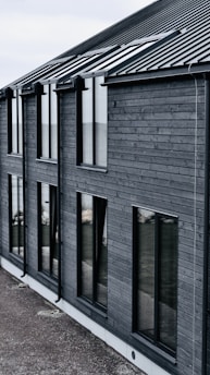 A modern building with dark wooden cladding and large rectangular windows. The roof is sloped and made of dark metal. The overall design is simple and minimalist.