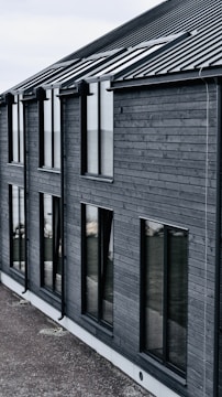 A modern building with dark wooden cladding and large rectangular windows. The roof is sloped and made of dark metal. The overall design is simple and minimalist.
