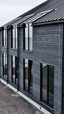 A modern building with dark wooden cladding and large rectangular windows. The roof is sloped and made of dark metal. The overall design is simple and minimalist.