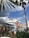 A luxurious yacht is docked by a picturesque marina, surrounded by lush tropical foliage and towering palm trees. The backdrop features an elaborate, castle-like pink structure with spires, reflecting a grand architectural design. The sky is partly cloudy with a mix of gray and blue hues.