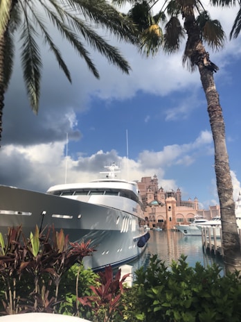 A luxurious yacht is docked by a picturesque marina, surrounded by lush tropical foliage and towering palm trees. The backdrop features an elaborate, castle-like pink structure with spires, reflecting a grand architectural design. The sky is partly cloudy with a mix of gray and blue hues.