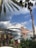 A luxurious yacht is docked by a picturesque marina, surrounded by lush tropical foliage and towering palm trees. The backdrop features an elaborate, castle-like pink structure with spires, reflecting a grand architectural design. The sky is partly cloudy with a mix of gray and blue hues.