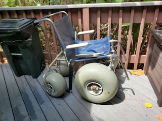 A wheelchair with large pneumatic wheels is positioned on a wooden deck next to a black garbage can. The wheelchair has a blue seat and backrest with gray metal frame. The deck is surrounded by a wooden fence, and there are leaves on the ground.