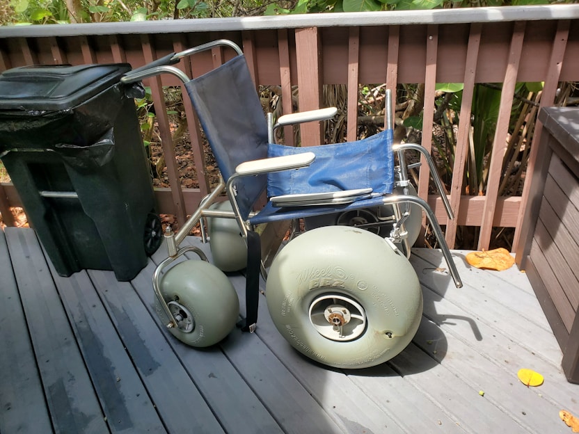A wheelchair with large pneumatic wheels is positioned on a wooden deck next to a black garbage can. The wheelchair has a blue seat and backrest with gray metal frame. The deck is surrounded by a wooden fence, and there are leaves on the ground.