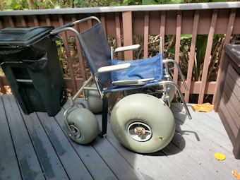 A wheelchair with large pneumatic wheels is positioned on a wooden deck next to a black garbage can. The wheelchair has a blue seat and backrest with gray metal frame. The deck is surrounded by a wooden fence, and there are leaves on the ground.