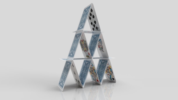 A structure composed of playing cards arranged in a triangular formation, resembling a house of cards. The cards feature various designs and suits, including spades and face cards, standing on a reflective surface against a plain background.