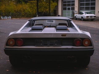 A vintage sports car is parked in an outdoor area with a second classic car visible in the background. The main car has a sleek design with a black bumper and dual exhausts, and the badge on the rear suggests it is a classic model. Autumn leaves are scattered on the ground.