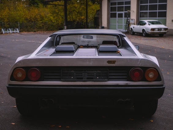 A vintage sports car is parked in an outdoor area with a second classic car visible in the background. The main car has a sleek design with a black bumper and dual exhausts, and the badge on the rear suggests it is a classic model. Autumn leaves are scattered on the ground.