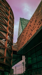 A modern architectural scene featuring a round building with grid-like patterns on the left, a triangular glass structure in the center, and a building with a brick-like facade on the right. The sky has a purple hue, creating a striking backdrop.