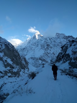A snowy mountain path leads towards towering, rugged peaks under a bright, clear sky. A solitary figure walks along the snow-covered trail, surrounded by steep rocky slopes.