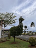 A creatively pruned tree shaped like an animal, possibly a bird, stands on a well-maintained lawn. The surrounding environment features other small trees, a lamp post, a road with a black and white curb, and a sky filled with clouds.