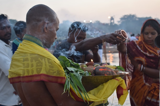 A group of people are gathered by the river, engaging in a ritual. One person is holding a metal tray filled with offerings, including green leaves and fruits, with incense sticks emitting smoke. The atmosphere is calm and reverent, and a woman in a colorful saree is pouring water as part of the ritual.