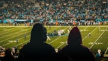 A football game is being played in a large stadium filled with spectators. Two people in hooded raincoats are in the foreground, observing the match. The field is brightly lit, and players in uniforms are lined up in formation. The crowd is dense, indicating a well-attended event.