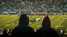 A football game is being played in a large stadium filled with spectators. Two people in hooded raincoats are in the foreground, observing the match. The field is brightly lit, and players in uniforms are lined up in formation. The crowd is dense, indicating a well-attended event.