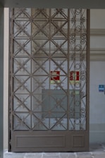A decorative metal gate featuring an intricate geometric pattern is set within an entranceway. Behind the gate, there are signs indicating directions such as 'Exit' in red on the wall. The surrounding walls are light-colored, with a tiled surface visible.