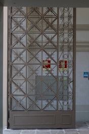 A decorative metal gate featuring an intricate geometric pattern is set within an entranceway. Behind the gate, there are signs indicating directions such as 'Exit' in red on the wall. The surrounding walls are light-colored, with a tiled surface visible.