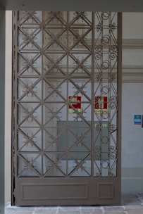 A decorative metal gate featuring an intricate geometric pattern is set within an entranceway. Behind the gate, there are signs indicating directions such as 'Exit' in red on the wall. The surrounding walls are light-colored, with a tiled surface visible.