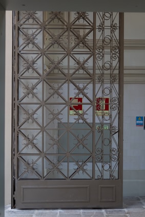 A decorative metal gate featuring an intricate geometric pattern is set within an entranceway. Behind the gate, there are signs indicating directions such as 'Exit' in red on the wall. The surrounding walls are light-colored, with a tiled surface visible.