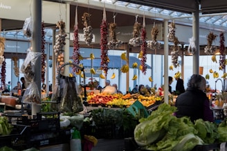 A bustling indoor market featuring a wide array of fresh produce including hanging garlic, chili peppers, and bananas. There are various fruits and vegetables displayed on tables, with customers browsing and engaging in conversation. A woman is visible in the foreground wearing a dark jacket.