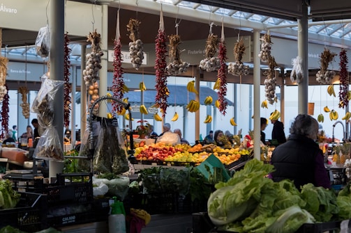 A bustling indoor market featuring a wide array of fresh produce including hanging garlic, chili peppers, and bananas. There are various fruits and vegetables displayed on tables, with customers browsing and engaging in conversation. A woman is visible in the foreground wearing a dark jacket.