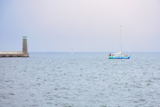 A sailboat with the words 'Ocean Team' on its side is moving across a calm sea. A lighthouse stands on a pier to the left, with a slightly overcast sky in the background.