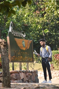 A person with a backpack and a gadget in hand stands next to a large wooden sign reading 'Welcome' amidst a lush, green park setting. The person appears to be observing something on the gadget. The scene is framed with various trees and bushes, creating a natural and peaceful environment.