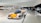 A sleek car with a Shell logo is prominently displayed in a modern showroom. Other cars are visible in the background, along with a group of people gathered near a white car marked 'Polizei'. The showroom features a futuristic design with white walls and floors.