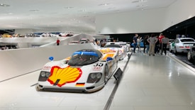 A sleek car with a Shell logo is prominently displayed in a modern showroom. Other cars are visible in the background, along with a group of people gathered near a white car marked 'Polizei'. The showroom features a futuristic design with white walls and floors.